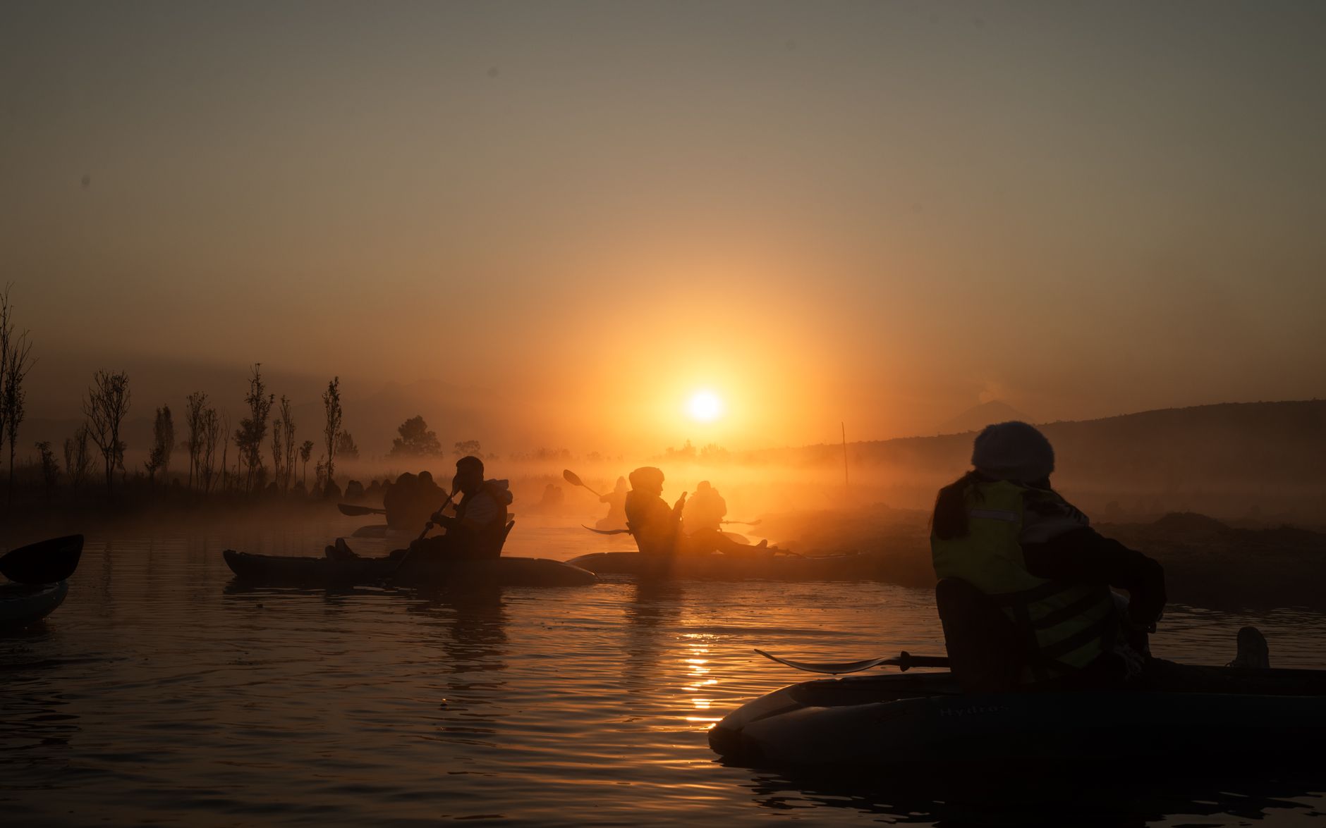 Silhouetted kayakers paddle through morning mist at sunrise on a tranquil lake.